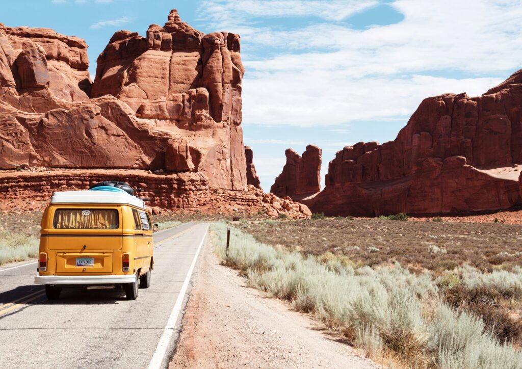 An orange VW van drives on a dirt road in a desert with red rock formations in the distance.