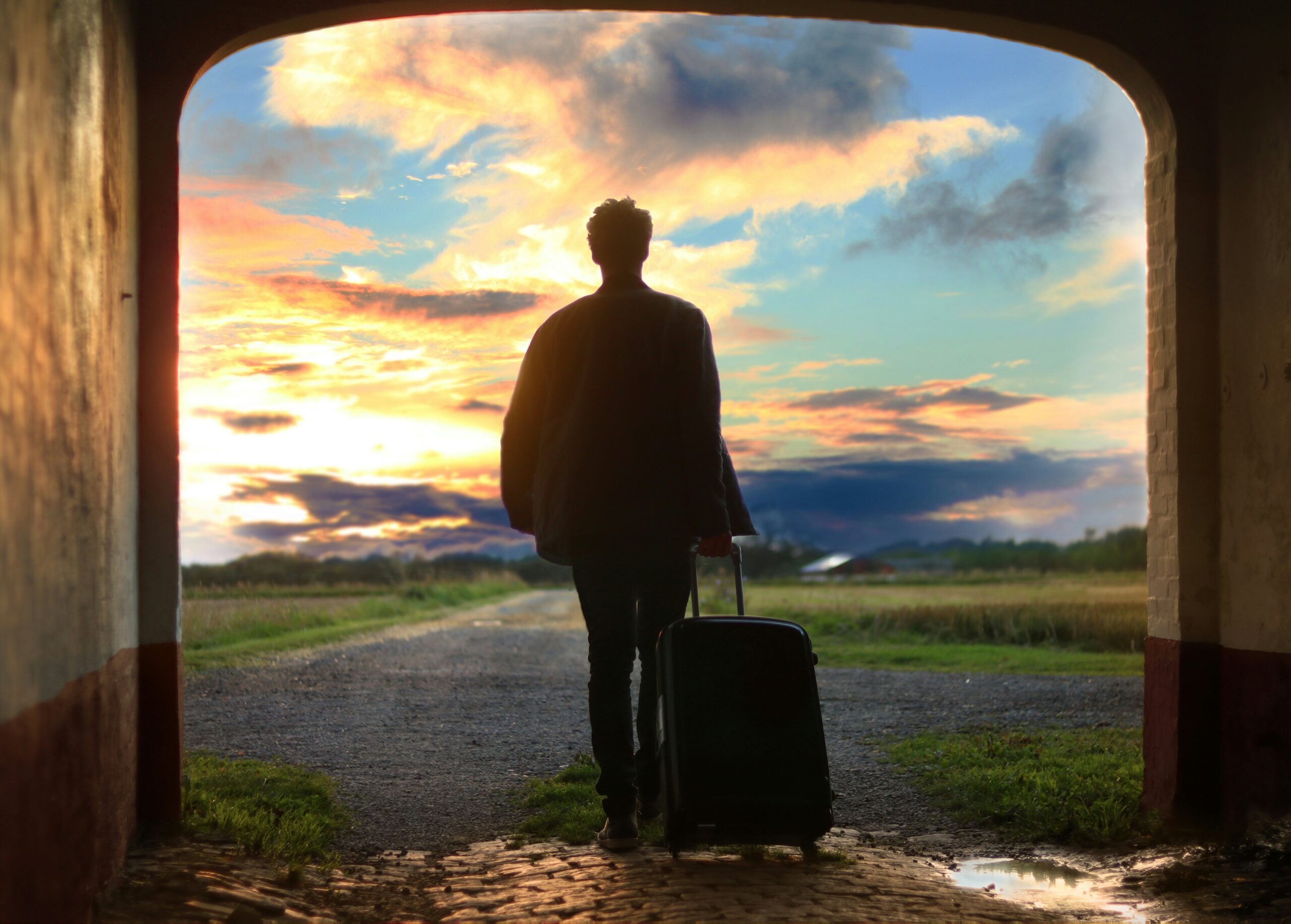 A traveler stands in an archway, pulling a wheeled suitcase, stepping into a landscape with a sunset.