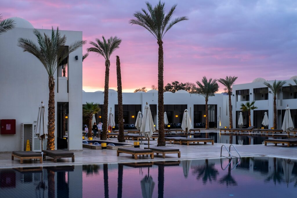 A white hotel surrounded by palm trees, with a sunset in the background, reflected in the pool in the foreground.