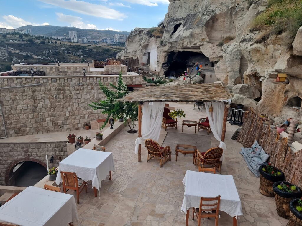 A cliffside with restaurant tables and chairs on a stone patio, overlooking a mountainous view.